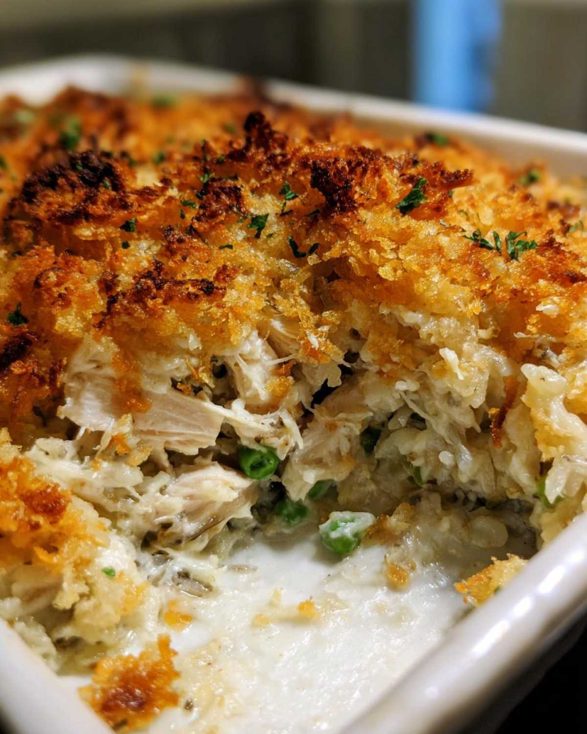 A scoop taken out of a creamy chicken wild rice casserole revealing shredded chicken, rice, peas, and a golden, crunchy breadcrumb topping.