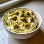 A white bowl filled with creamy dirty martini dip, topped generously with green olives and a drizzle of olive oil.