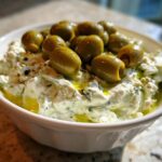 Close-up of a creamy, white dirty martini dip in a bowl, generously topped with whole green olives and olive oil.