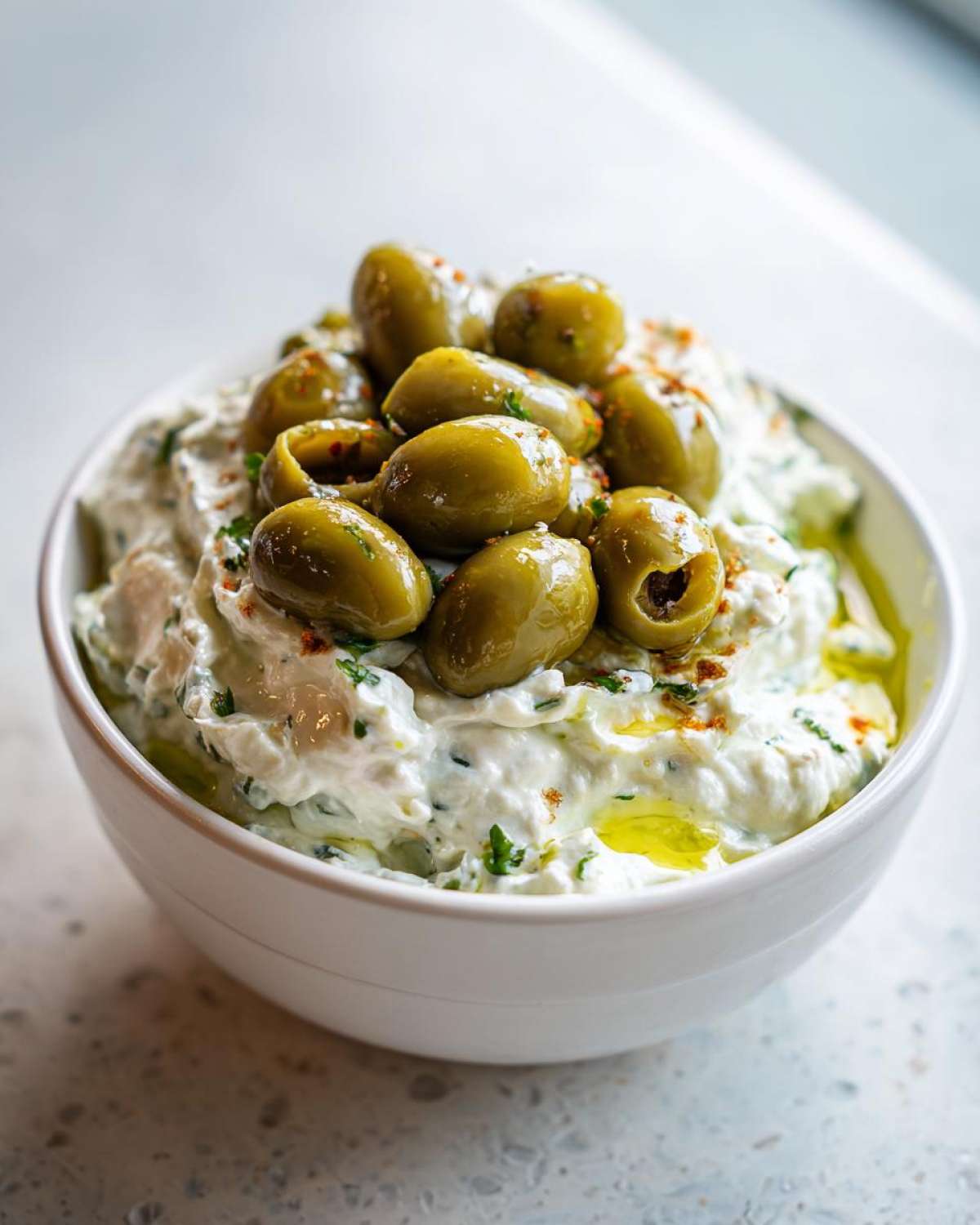 Close-up of a creamy white dirty martini dip garnished generously with whole green olives and a drizzle of olive oil.