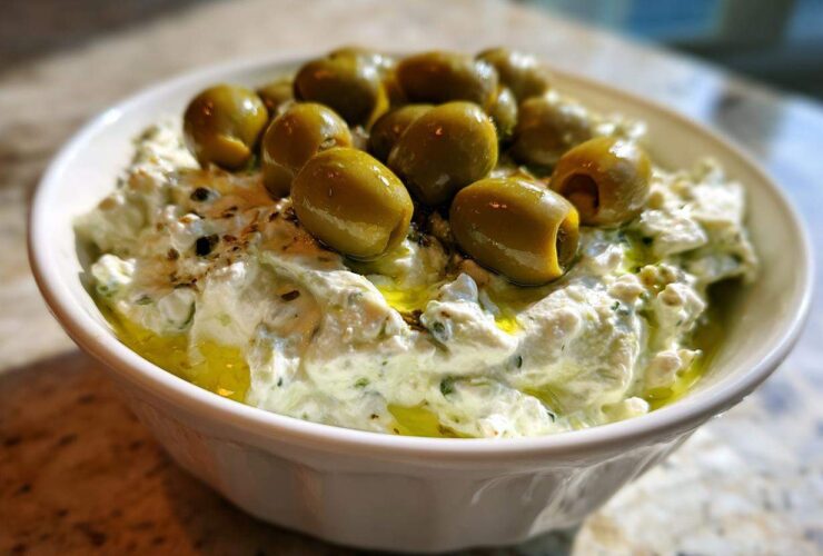 Close-up of a creamy, white dirty martini dip in a bowl, generously topped with whole green olives and olive oil.