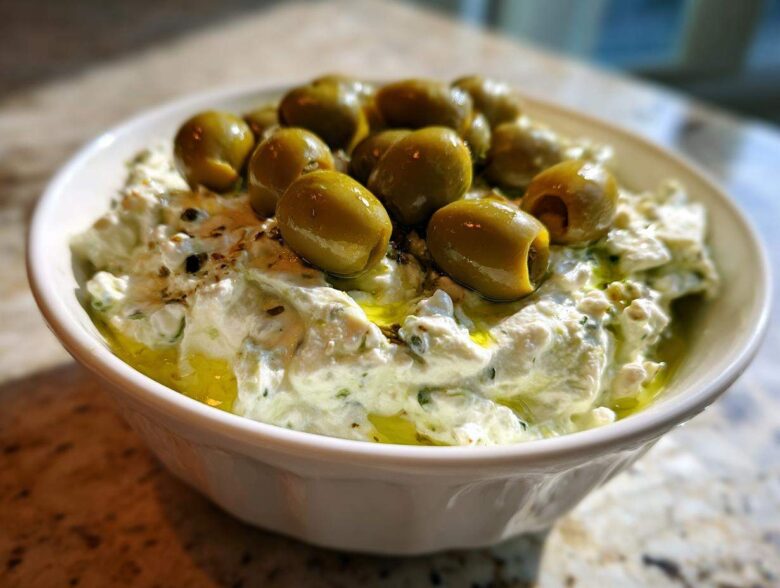 Close-up of a creamy, white dirty martini dip in a bowl, generously topped with whole green olives and olive oil.