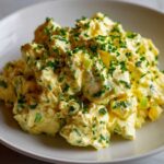 A close-up of creamy, yellow egg salad recipe generously topped with chopped green chives and celery.