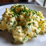A mound of creamy, yellow egg salad recipe garnished generously with freshly chopped green chives on a white plate.