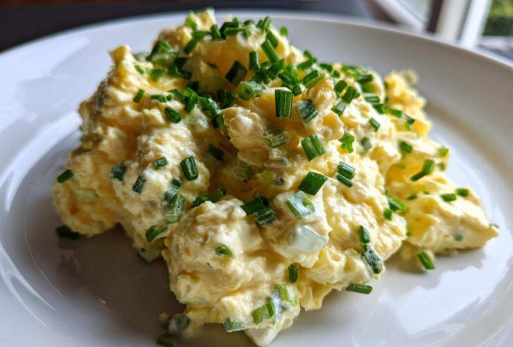 A mound of creamy, yellow egg salad recipe garnished generously with freshly chopped green chives on a white plate.