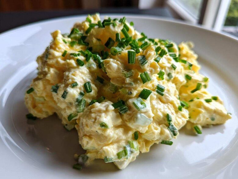 A mound of creamy, yellow egg salad recipe garnished generously with freshly chopped green chives on a white plate.