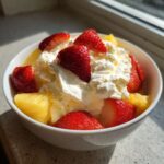 A white bowl filled with chunks of pineapple, sliced strawberries, and topped generously with whipped cream, likely served with a fruit dip.