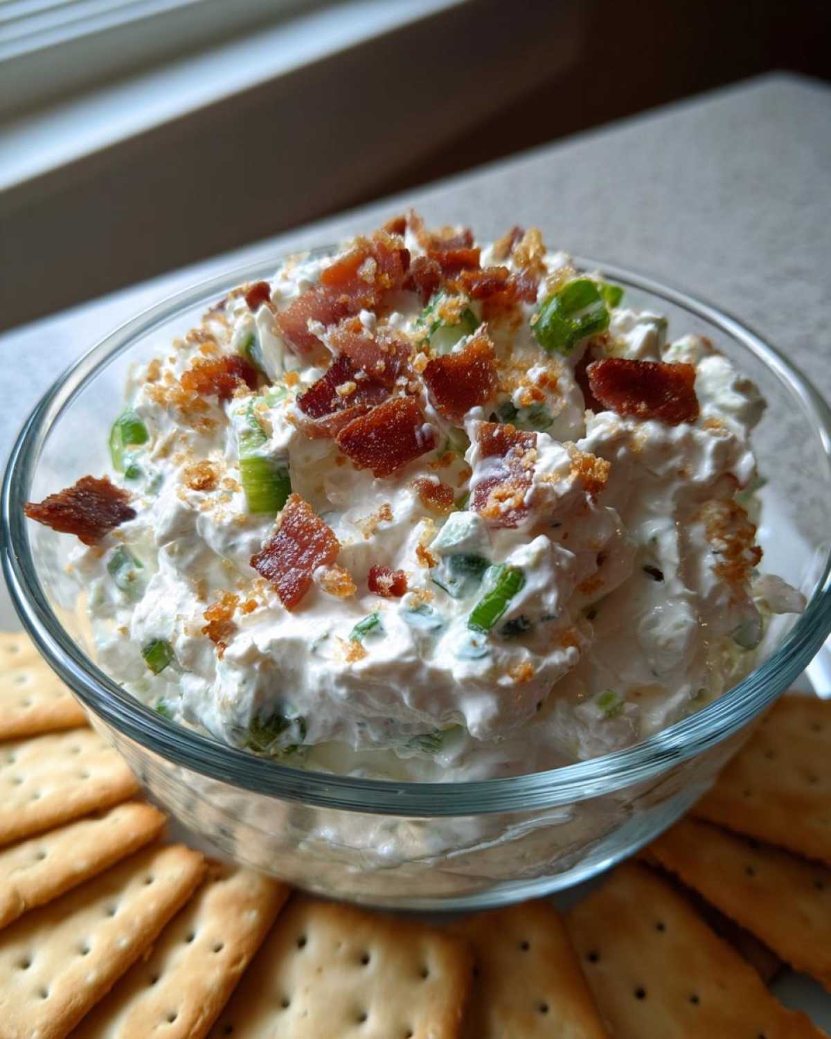 A close-up of creamy Million Dollar Dip in a glass bowl, topped with crumbled bacon and green onions, served with crackers.