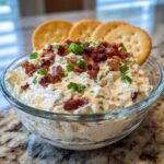 A glass bowl filled with creamy Million Dollar Dip, topped generously with crispy bacon bits and chopped green onions, served with crackers.