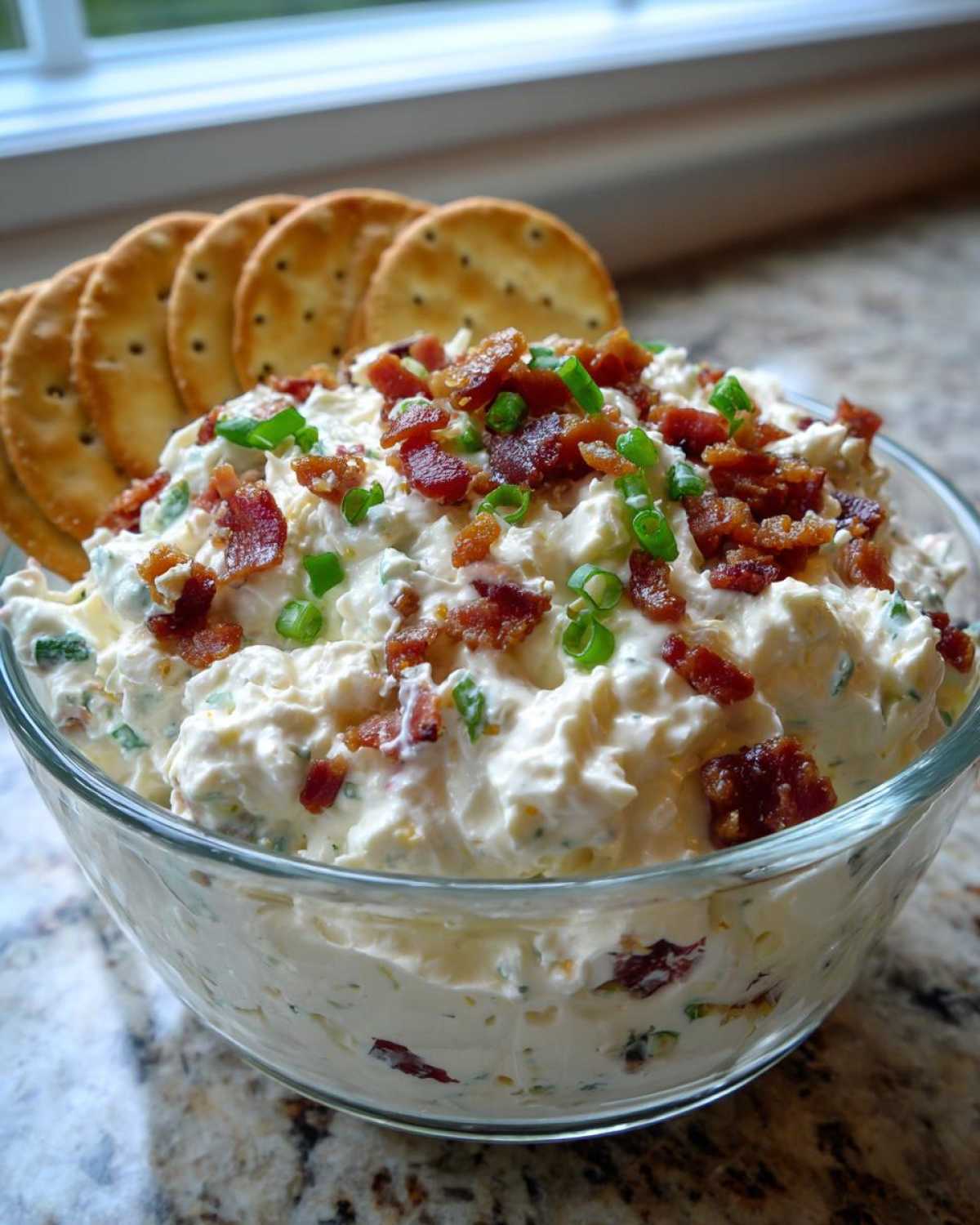 A bowl of creamy Million Dollar Dip topped generously with crispy bacon bits and fresh green onions, served with crackers.