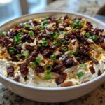 A white bowl filled with creamy Neiman Marcus Dip, heavily garnished with bacon bits, sliced almonds, and chopped green onions.