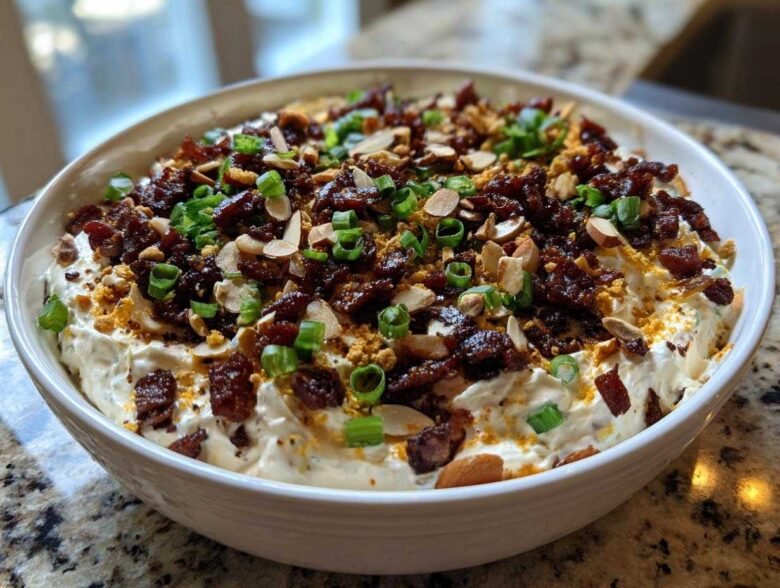 A white bowl filled with creamy Neiman Marcus Dip, heavily garnished with bacon bits, sliced almonds, and chopped green onions.