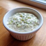 Close-up of creamy, chunky tartar sauce recipe served in a small white bowl on a wooden surface.