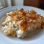 A close-up serving of creamy Tuna Casserole topped with golden, crispy potato chips or breadcrumbs, garnished with herbs.