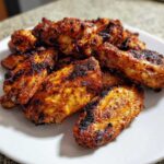 A close-up of a pile of golden brown, heavily seasoned, and crispy baked chicken wings on a white serving plate.