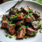 Close-up of tender crockpot steak bites glazed in sauce and topped with fresh chopped parsley on a white plate.