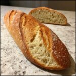 Close-up of a golden brown, crusty French bread loaf with an open crumb texture, sitting on a speckled countertop.