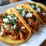Close-up of three soft shell chicken tacos filled with shredded seasoned chicken, topped with diced white onion and cilantro.