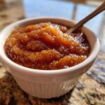 A close-up shot of rich, dark brown homemade applesauce served in a small white fluted ramekin with a spoon.