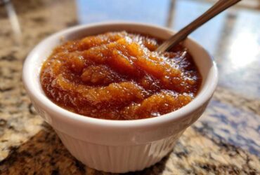 A close-up shot of rich, dark brown homemade applesauce served in a small white fluted ramekin with a spoon.