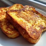 Close-up of two golden-brown slices of easy french toast stacked slightly on a white plate, backlit by sunlight.