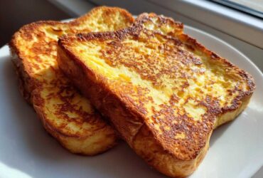 Close-up of two golden-brown slices of easy french toast stacked slightly on a white plate, backlit by sunlight.