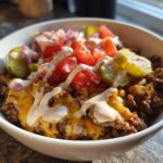 A close-up of a white bowl filled with a cheeseburger bowl featuring seasoned ground beef, melted cheese, pickles, tomatoes, onions, and a drizzle of sauce.