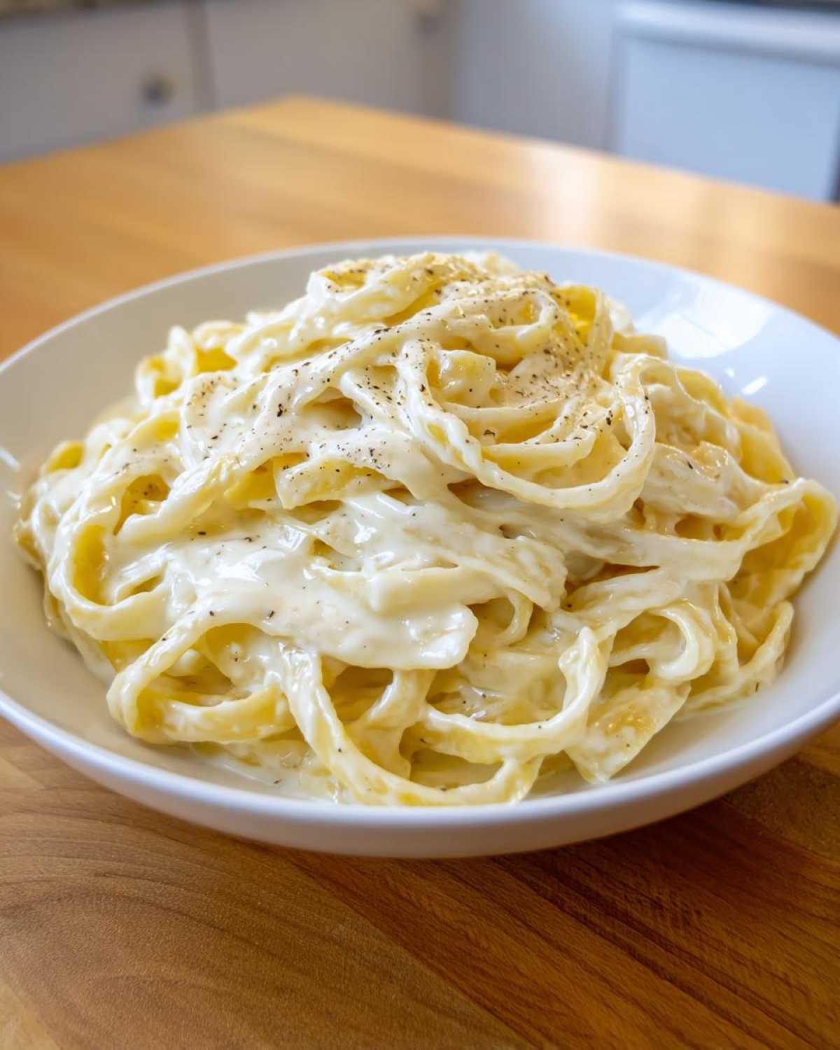 A white bowl filled with fettuccine pasta generously coated in creamy homemade alfredo sauce and topped with black pepper.