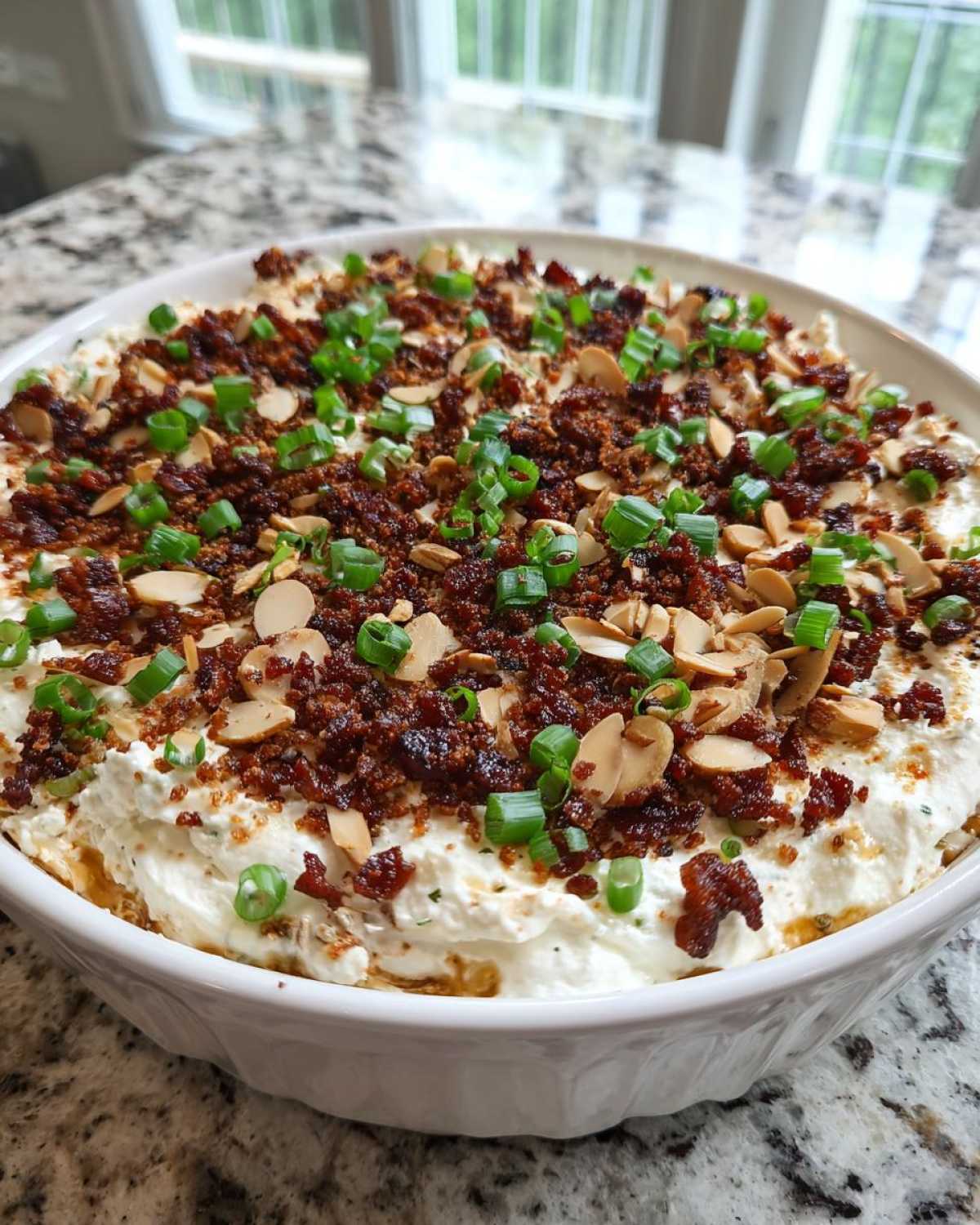 A creamy Neiman Marcus dip topped with crumbled bacon, sliced almonds, and green onions in a white serving bowl.