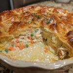 A close-up of a freshly baked turkey pot pie with a slice removed, showing creamy filling, turkey, carrots, and peas under a golden, flaky crust.