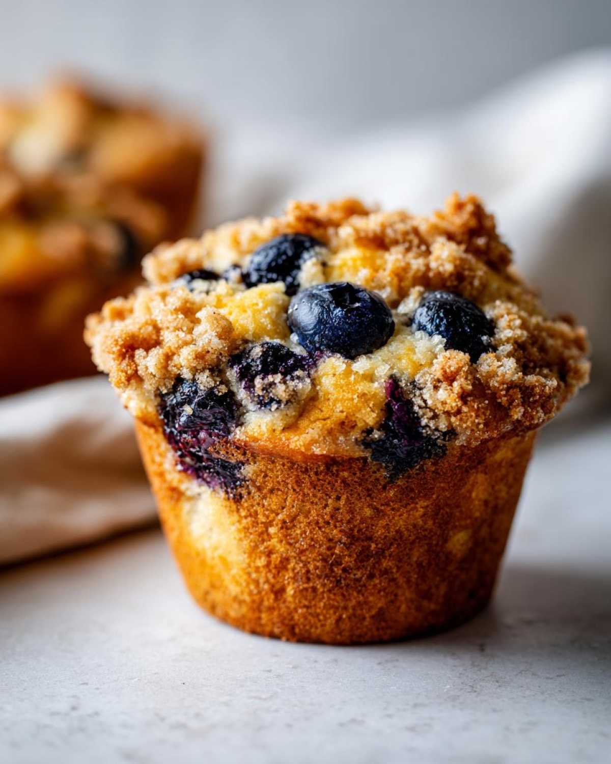 Close-up of a perfectly baked blueberry muffin featuring a golden brown base and a generous, sugary crumb topping studded with fresh blueberries.