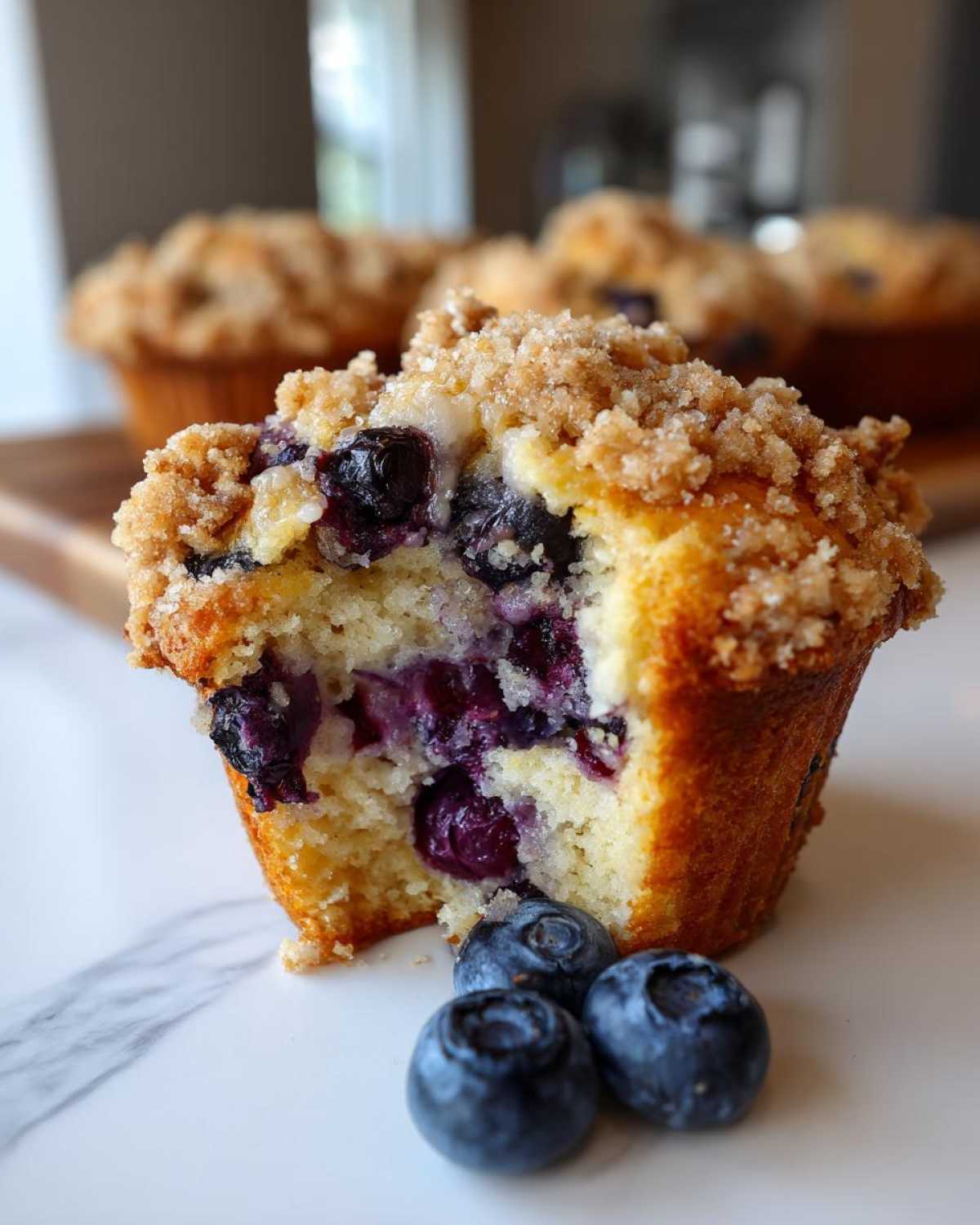 Close-up of a fluffy blueberry muffin cut in half, showing juicy berries and a streusel topping, with fresh blueberries nearby.