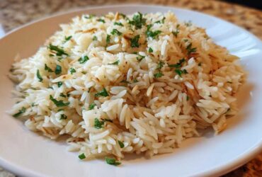 A close-up of fluffy rice pilaf, mixed with toasted vermicelli and garnished with fresh parsley, served on a white plate.