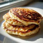 A close-up stack of three golden-brown, fluffy sourdough discard pancakes sitting on a white plate near a window.