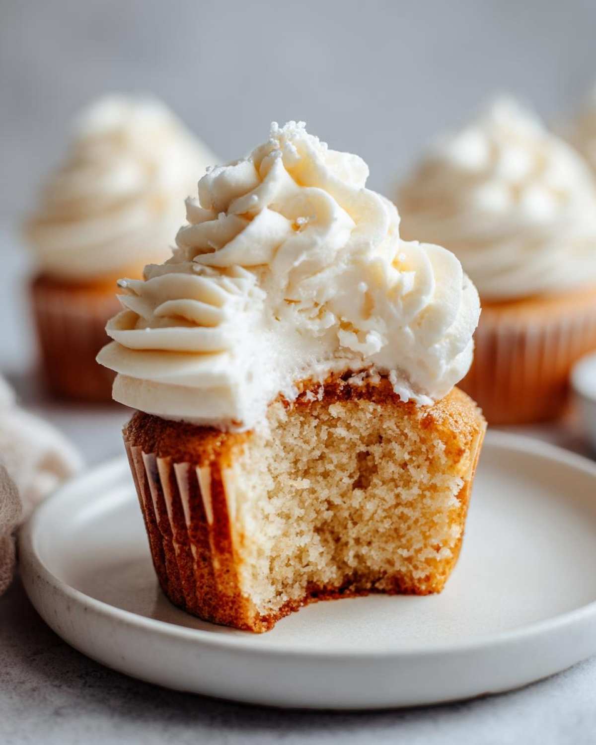 A vanilla cupcake with a bite taken out, showing the fluffy interior and generous swirl of white vanilla frosting.