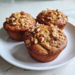 Close-up of three golden-brown banana nut muffins piled on a white plate, generously topped with chopped nuts.