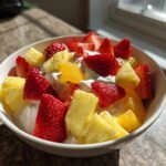 A white bowl filled with fresh strawberries, pineapple chunks, and orange segments served over a creamy fruit dip.