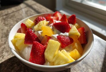A white bowl filled with fresh strawberries, pineapple chunks, and orange segments served over a creamy fruit dip.