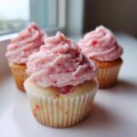 A close-up of one perfectly frosted strawberry cupcake, with two more strawberry cupcakes blurred in the background.