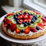 A vibrant fruit pizza sugar cookie base topped with whipped cream, sliced strawberries, kiwi, and blueberries.