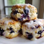 A stack of three fluffy, glazed blueberry scones bursting with fresh blueberries, served on a white plate.