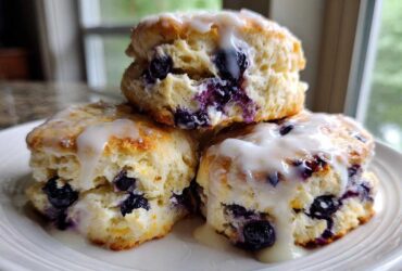 A stack of three fluffy, glazed blueberry scones bursting with fresh blueberries, served on a white plate.