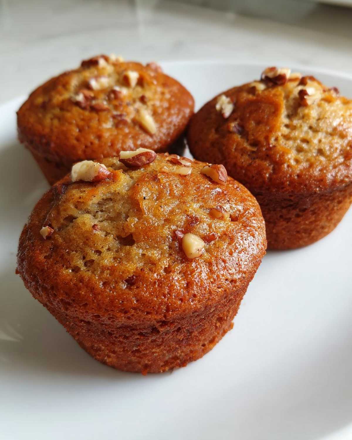 Three freshly baked banana nut muffins with a golden-brown, glossy top sprinkled with chopped pecans, resting on a white plate.