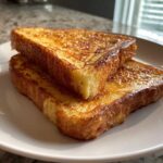 Two thick slices of golden brown french toast stacked slightly on a white plate.