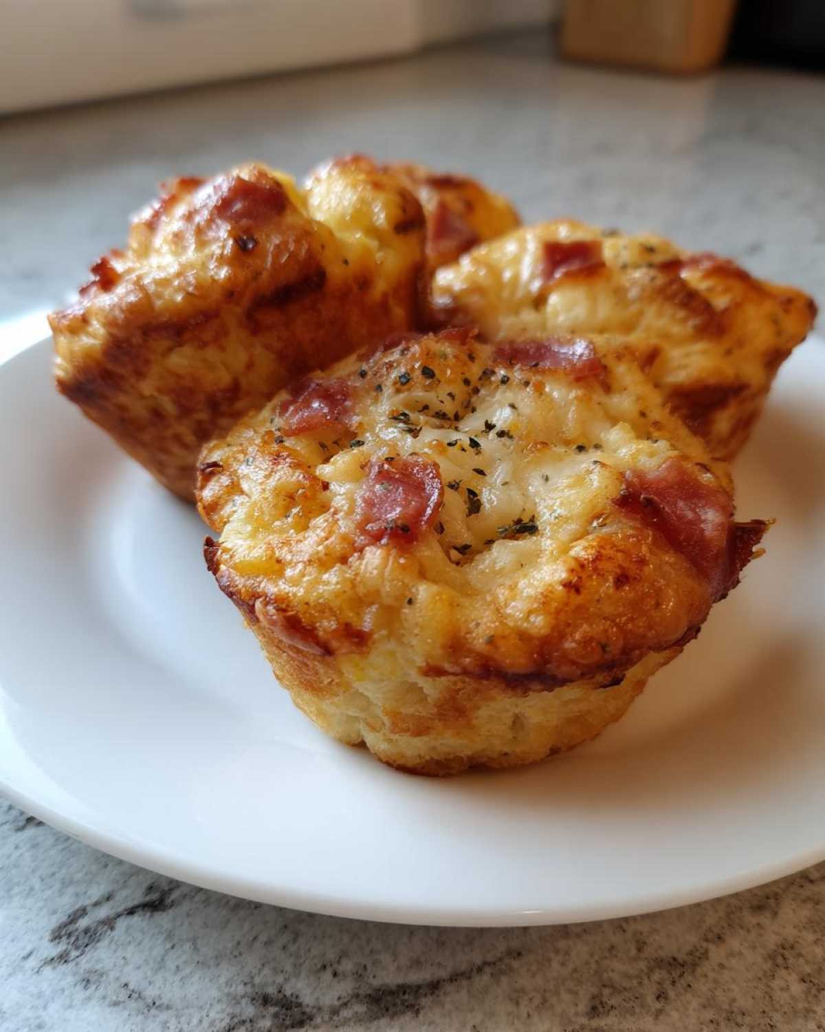 Three golden brown Pizza Muffins topped with melted cheese and pepperoni pieces, served on a white plate.