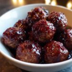 Close-up of several glistening Grape Jelly Meatballs coated in a rich, dark sauce, served in a white bowl.