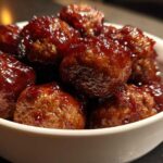 Close-up of shiny, glazed grape jelly meatballs piled high in a small white serving bowl.