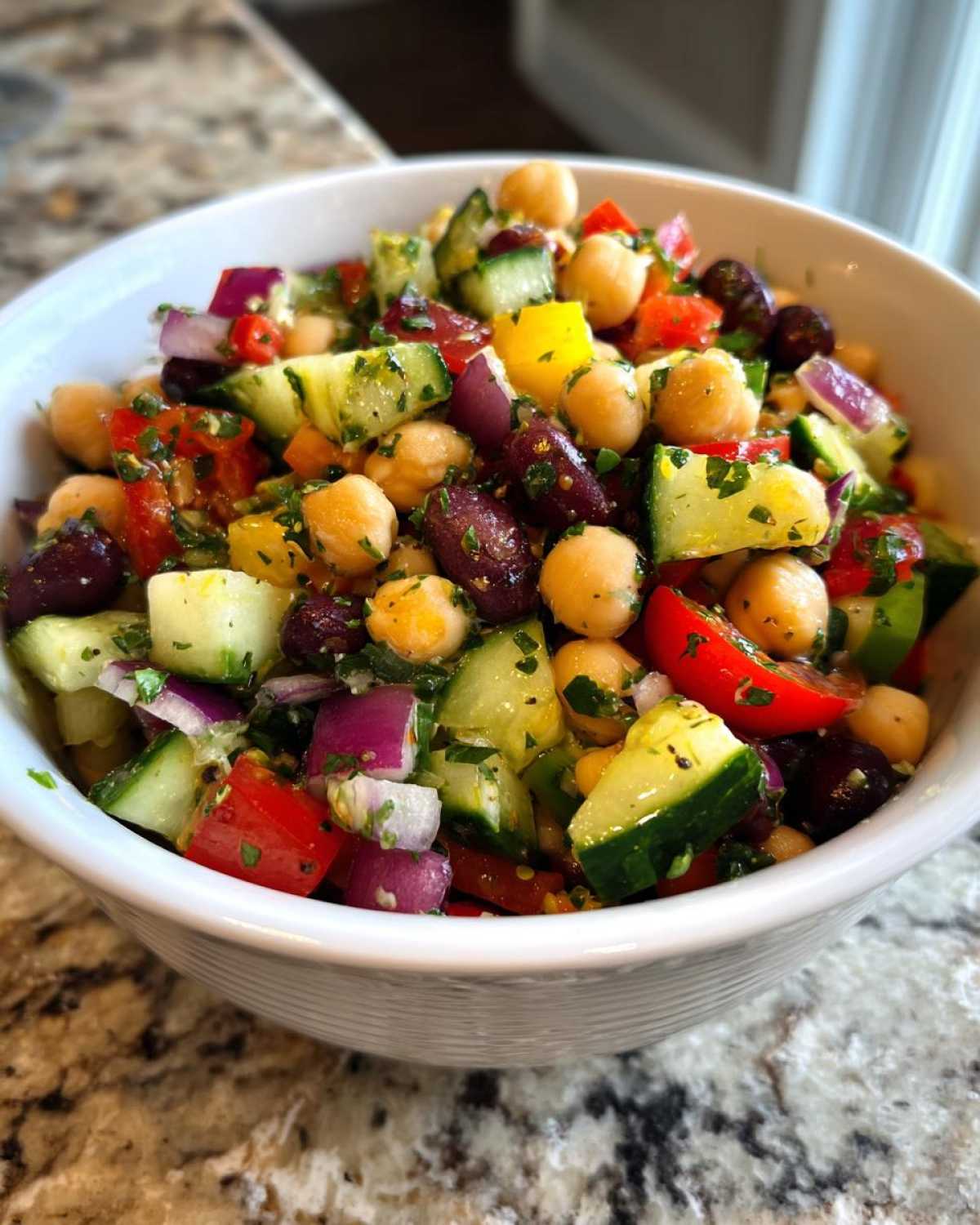 Close-up of a vibrant, colorful bean salad featuring chickpeas, kidney beans, cucumber, red onion, and tomatoes in a white bowl.