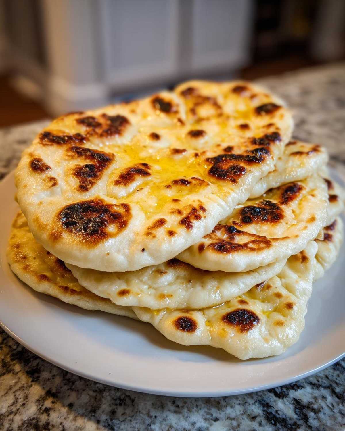 A stack of freshly made, buttery homemade naan bread with beautiful dark char marks, resting on a white plate.