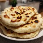 A close-up of a stack of freshly made homemade naan bread with golden-brown char marks on a white plate.